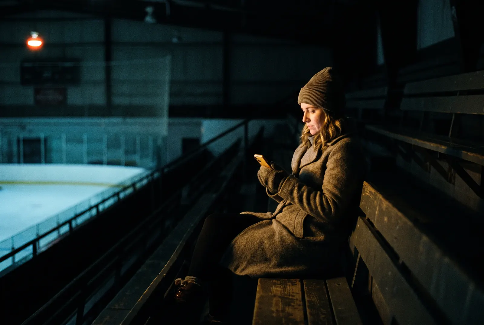 A parent reading a message on her phone alone in the dark stands of an ice rink.