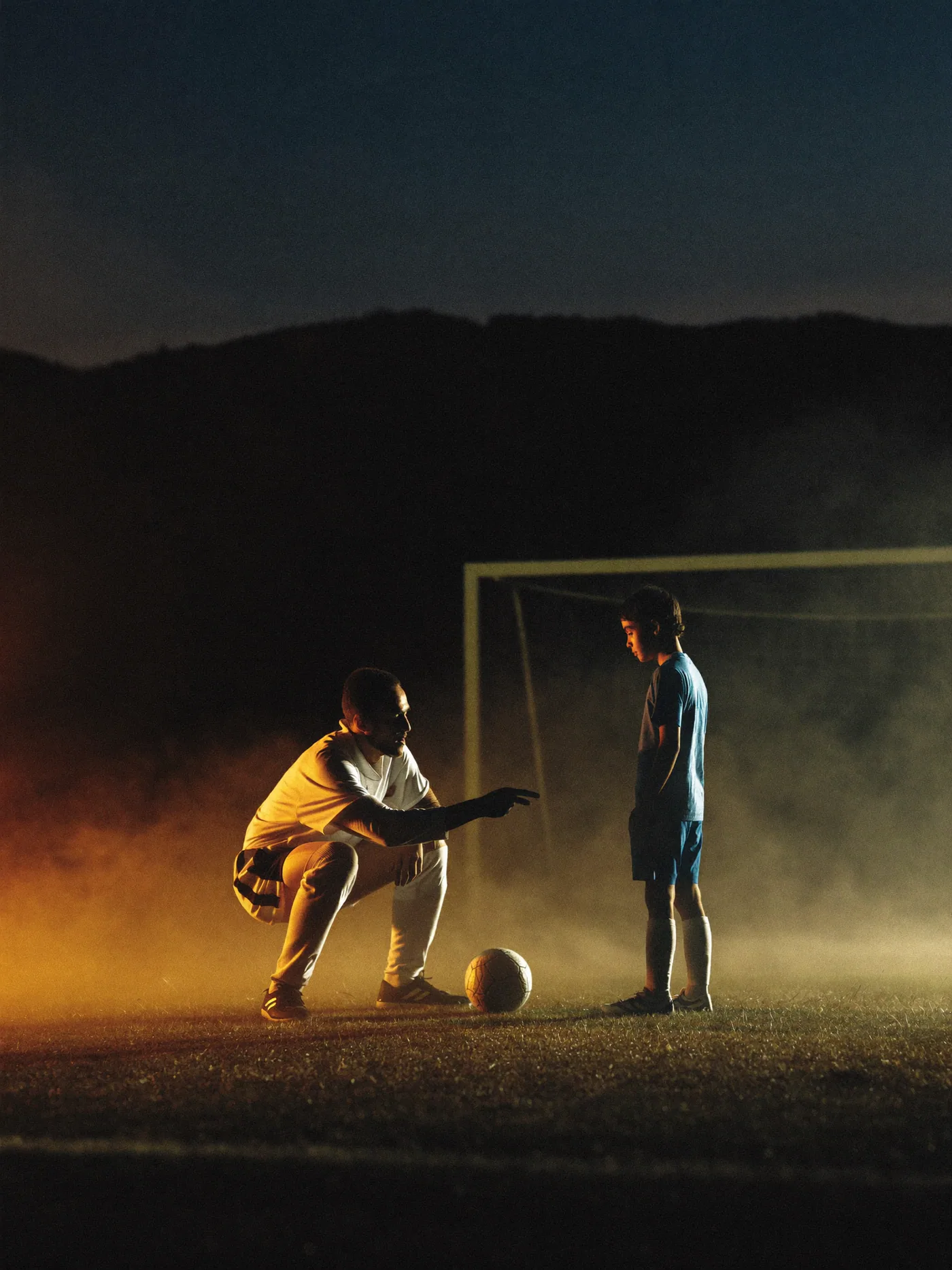 A youth soccer coach crouched to eye level, pointing out a play to a young player on a misty field.