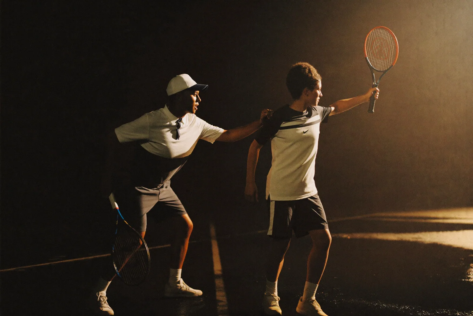 A coach steadying a young tennis player mid-rally under court floodlights.
