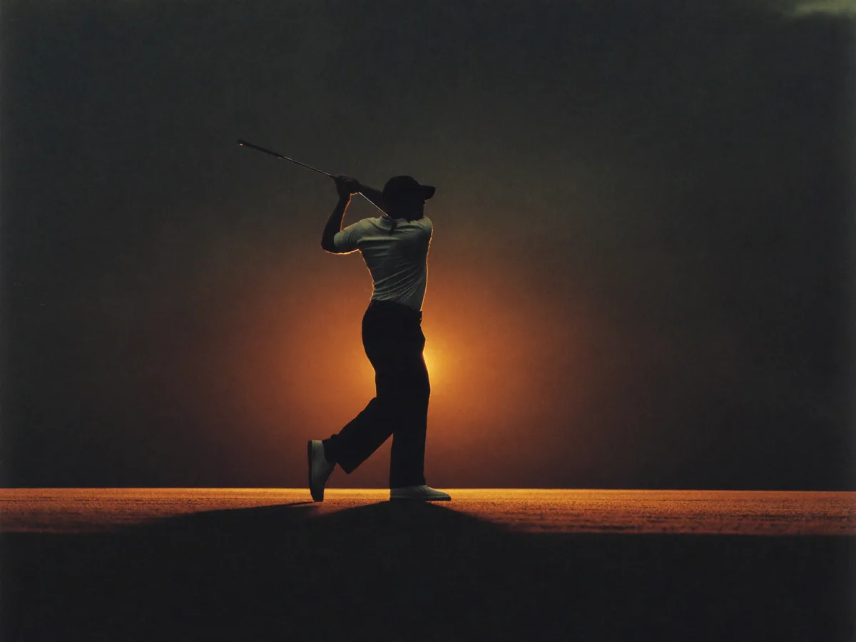 An adult golfer at the top of a swing, rim-lit against a dark course at dusk.