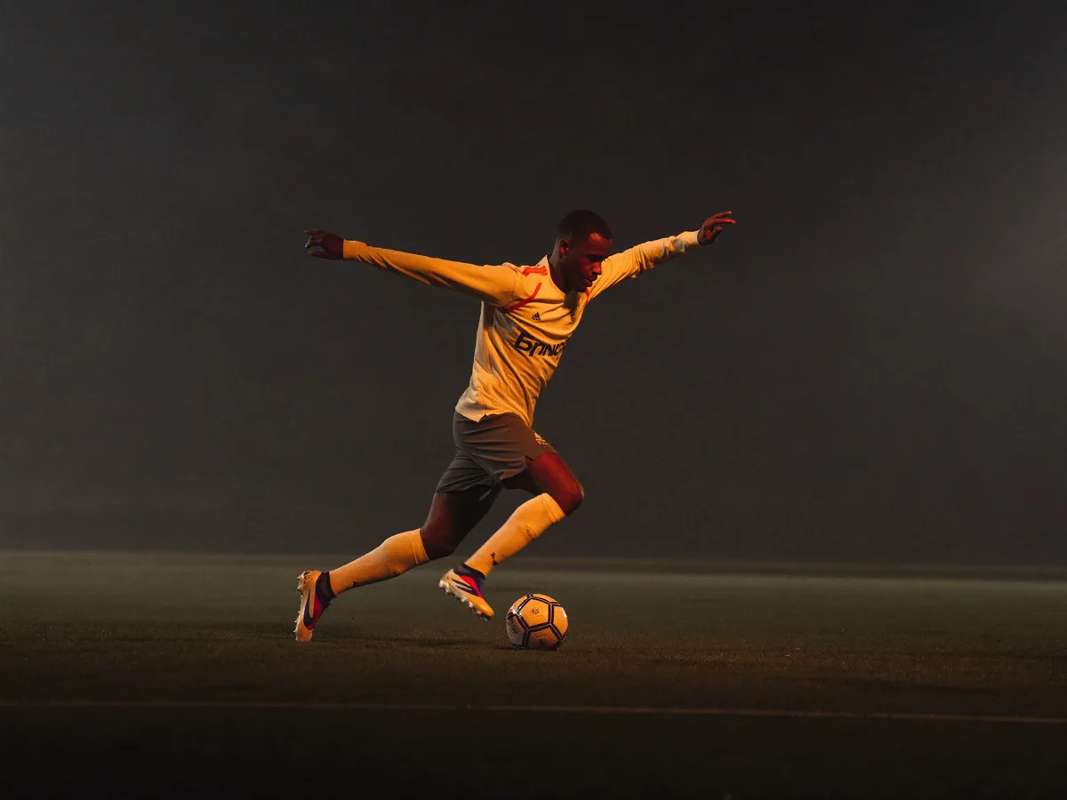 An adult soccer player striking the ball on a misty floodlit pitch at night.